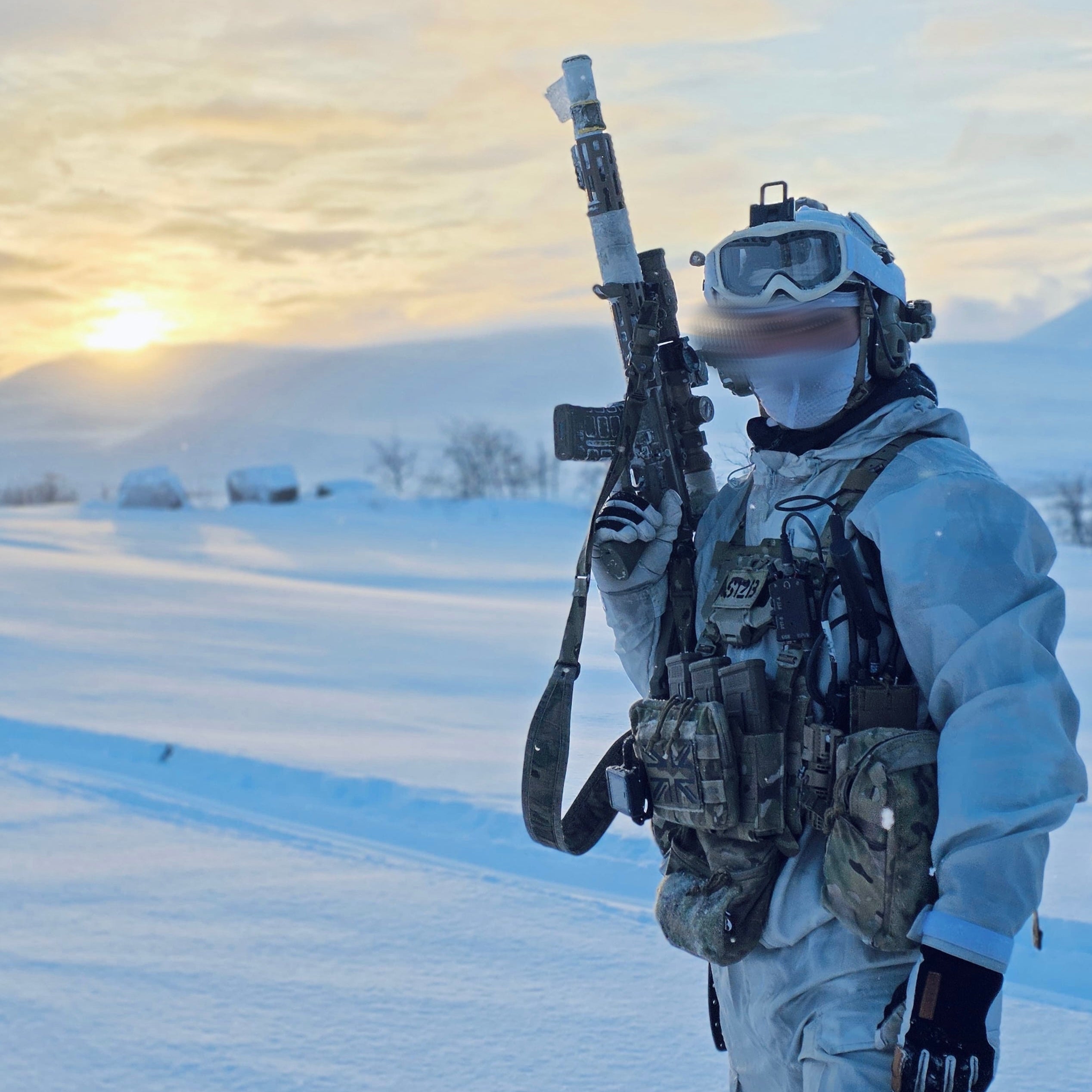 Person in military gear holding a rifle in a snowy landscape with a sunset.