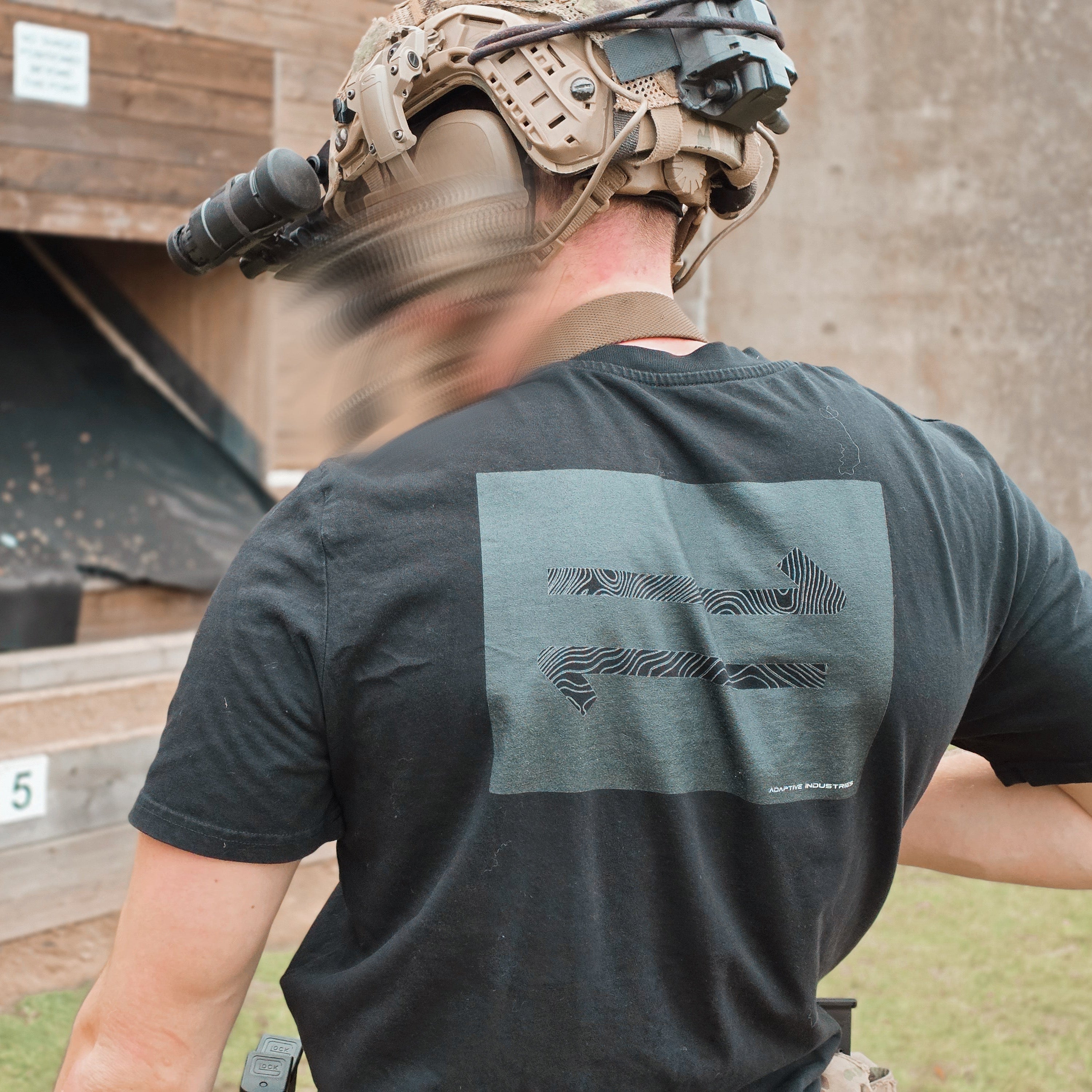 Person wearing tactical gear and a black t-shirt with a logo, standing outdoors.