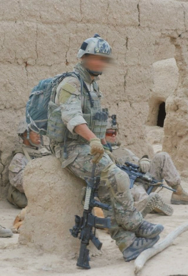 Soldier in camouflage sitting on a stone wall with a rifle, wearing a helmet and carrying a backpack.