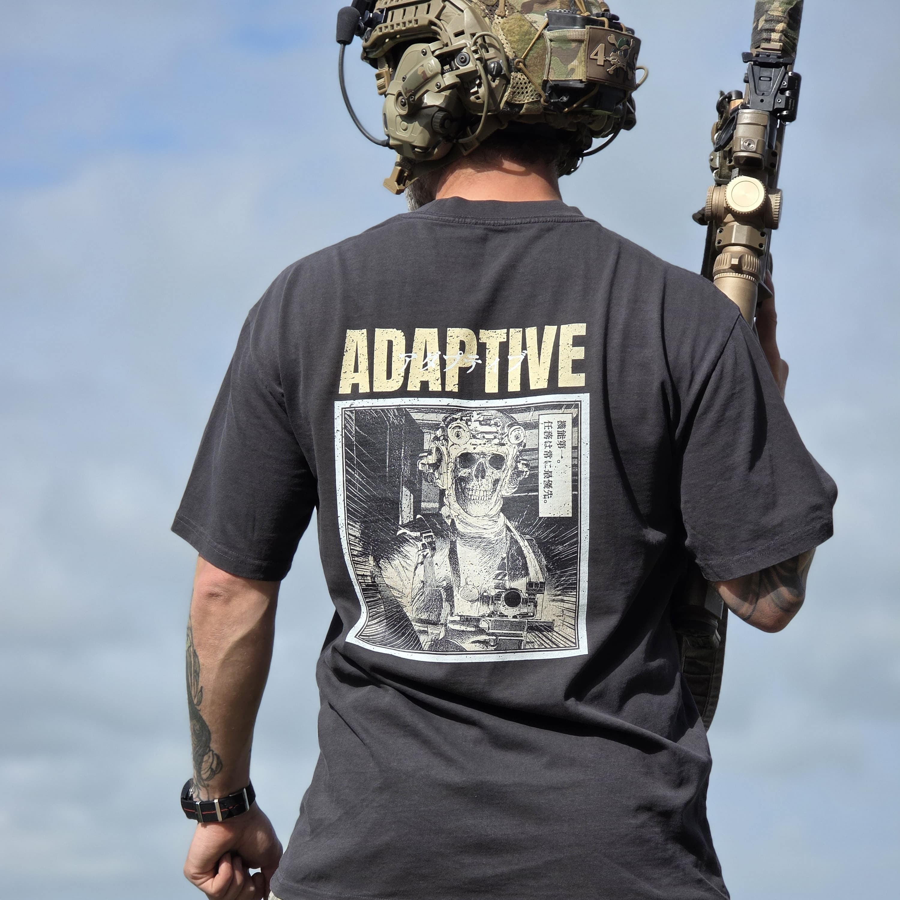 A man viewed from behind wearing a black Adaptive t-shirt with a large graphic print on the back, camouflage cargo pants, and a tactical helmet while standing outdoors.