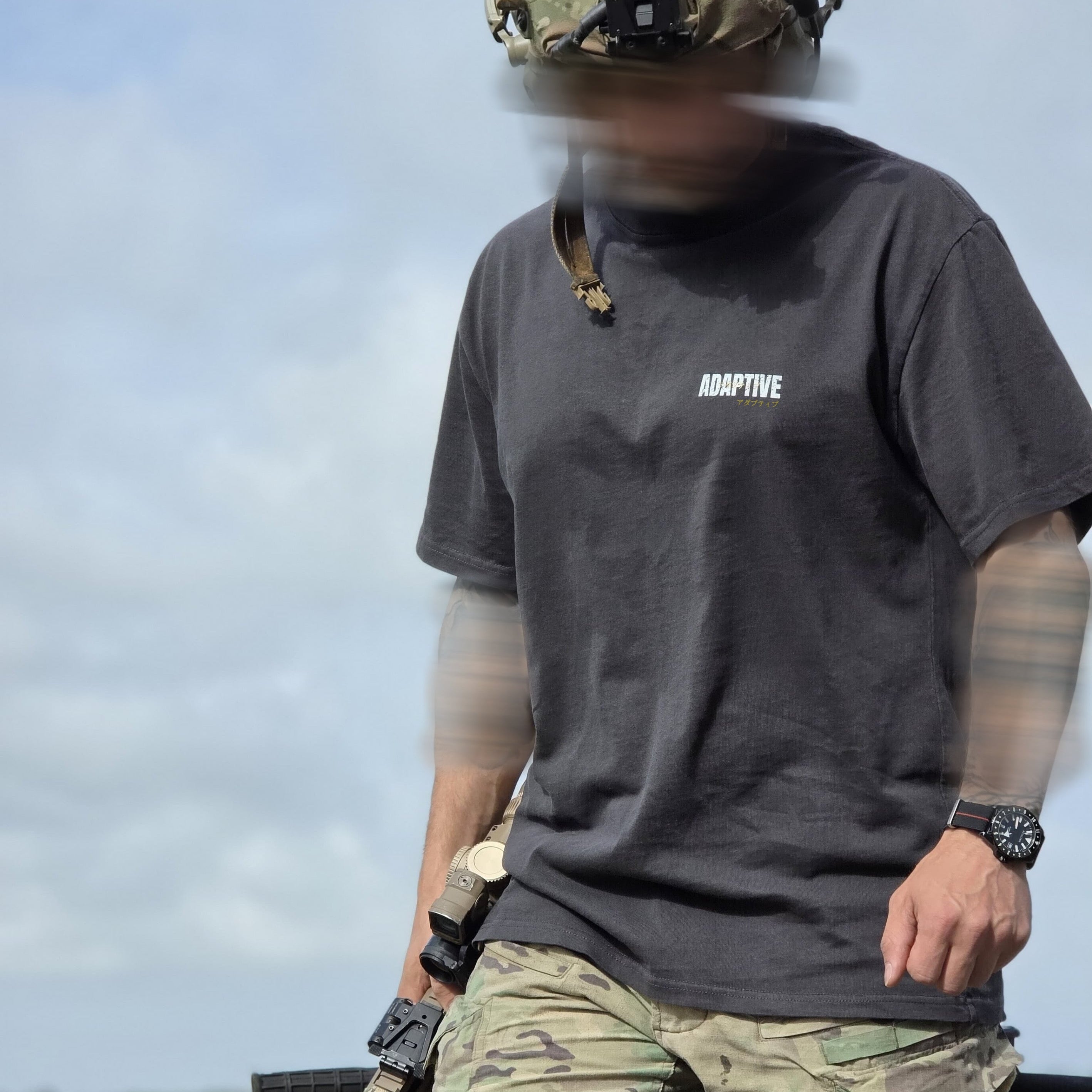 A person outdoors wearing a dark charcoal gray short-sleeve t-shirt with the word 'ADAPTIVE' printed in white on the left chest, paired with camouflage pants and a tactical helmet.