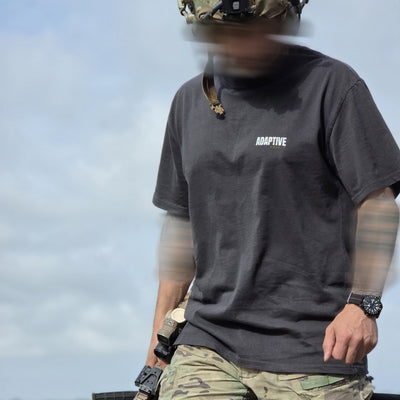 A person outdoors wearing a dark charcoal gray short-sleeve t-shirt with the word 'ADAPTIVE' printed in white on the left chest, paired with camouflage pants and a tactical helmet.