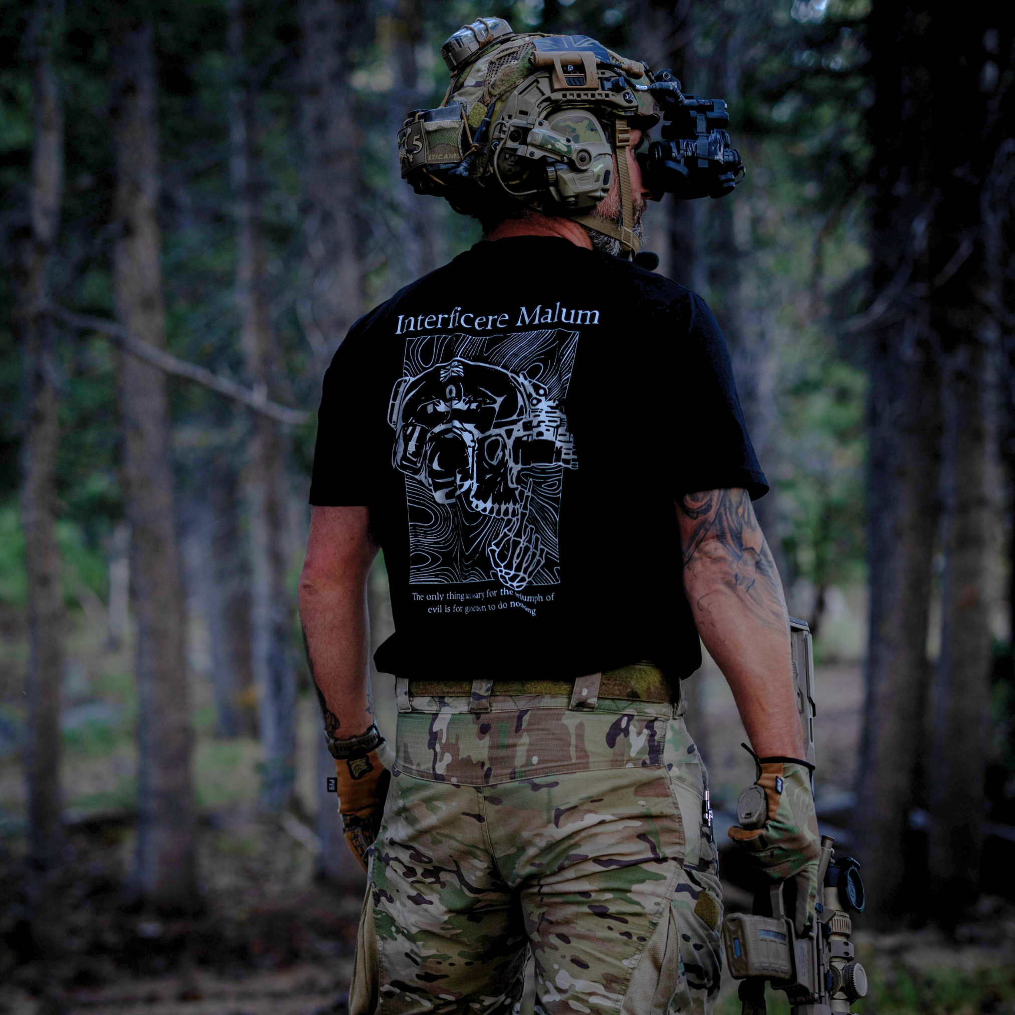 Person in tactical gear with a black t-shirt featuring a skull design in a forest setting